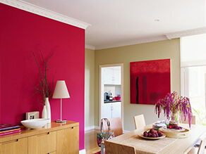 Bright pink feature wall in dining room with wooden table and red artwork plants and drawers